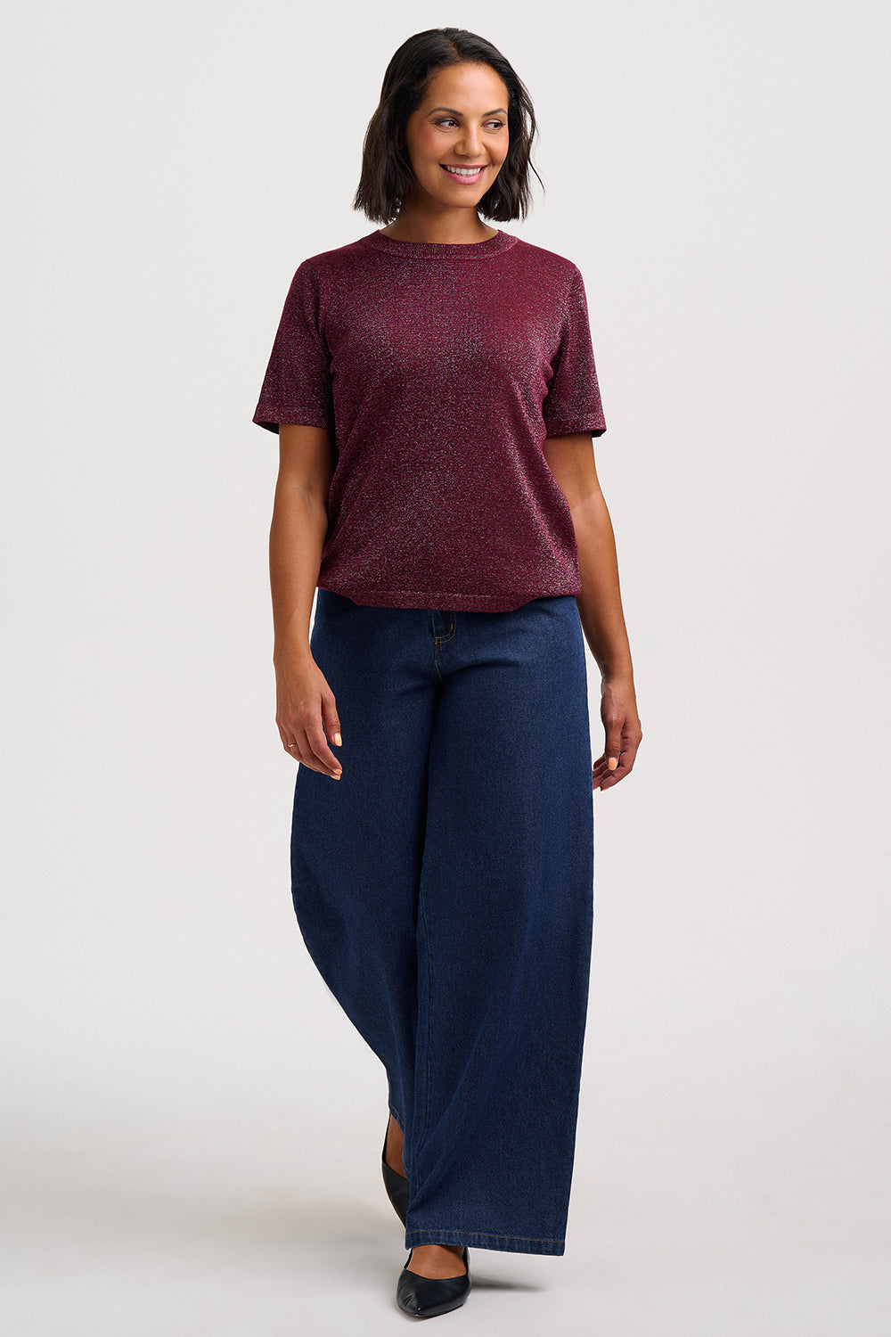 A woman modeling a short-sleeve crewneck top in a heathered burgundy color, paired with dark blue wide-leg jeans and black flats, against a plain light gray studio background.