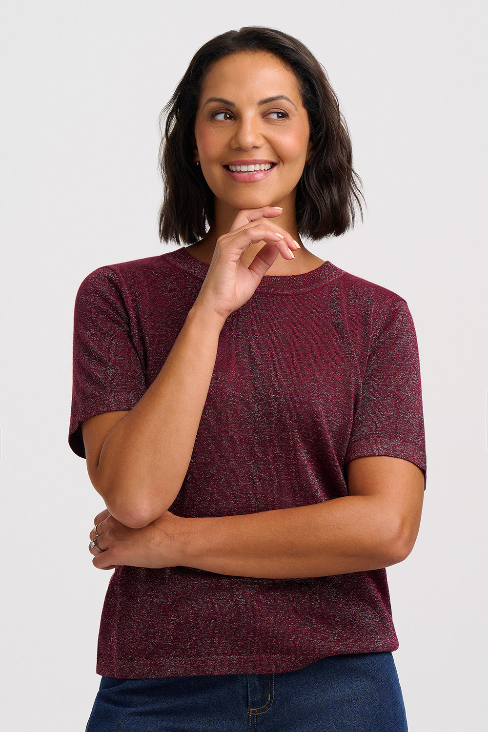 A woman with short dark hair wearing a marled burgundy short-sleeve crewneck t-shirt and blue jeans, posing against a light gray studio background.