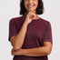 A woman with short dark hair wearing a marled burgundy short-sleeve crewneck t-shirt and blue jeans, posing against a light gray studio background.