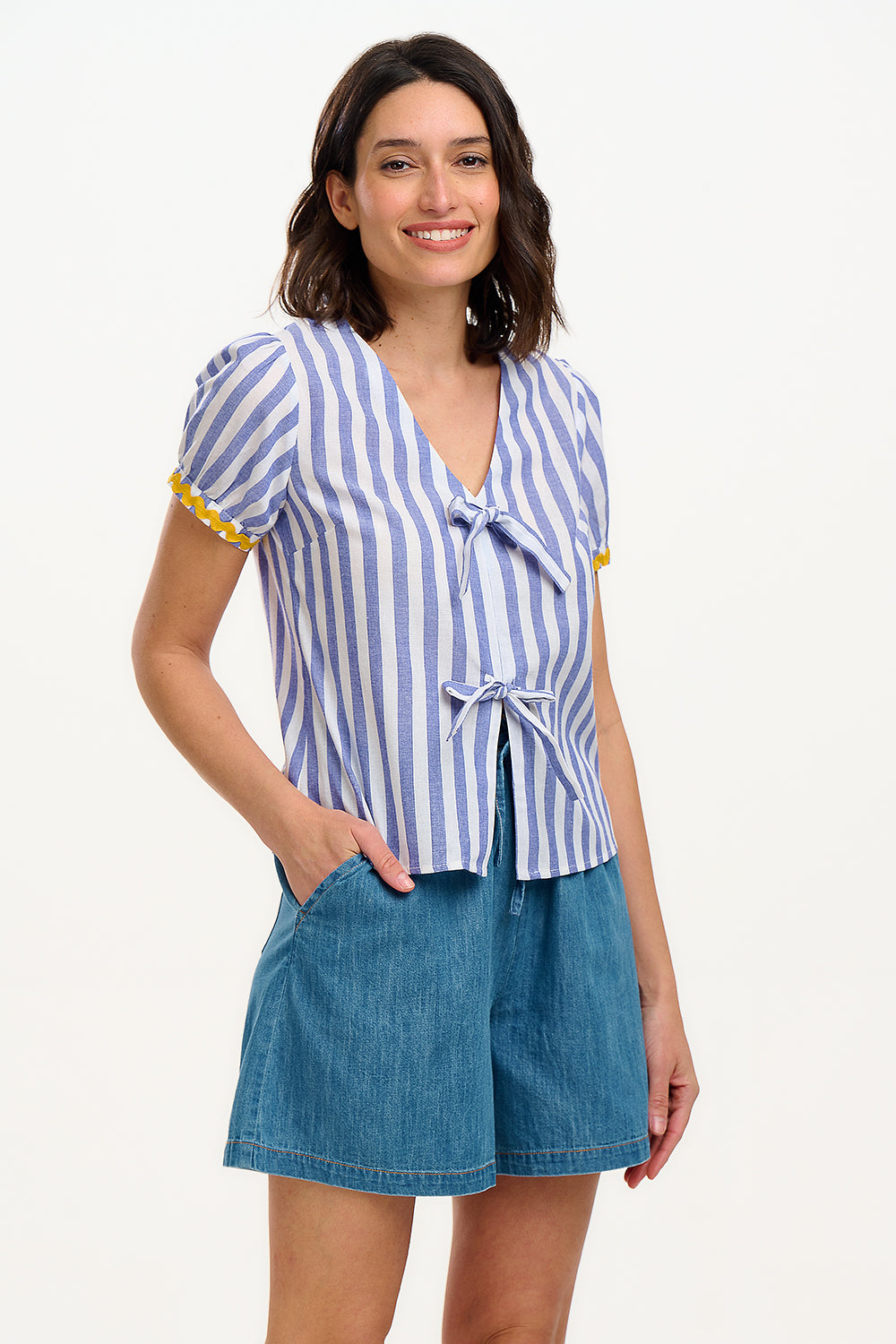 A woman smiling and wearing an off-white and blue striped top with short puff sleeves and a decorative bow tie at the v-neck.