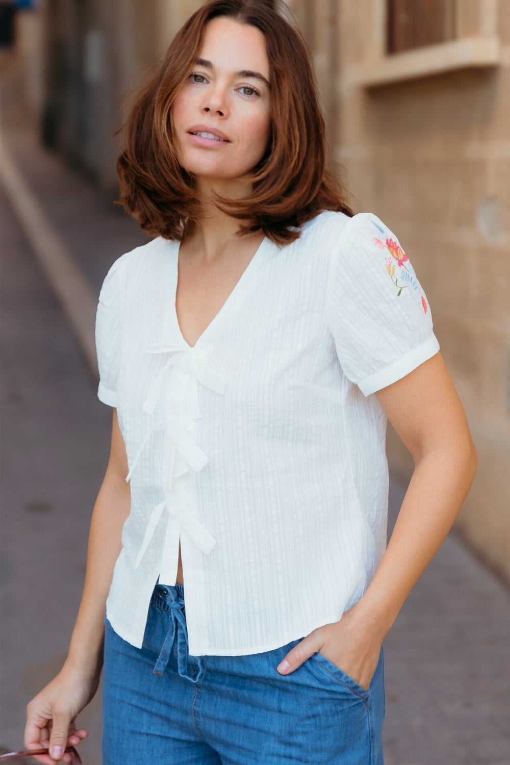 Woman wearing a white blouse with floral embroidery and blue jeans on a street.
