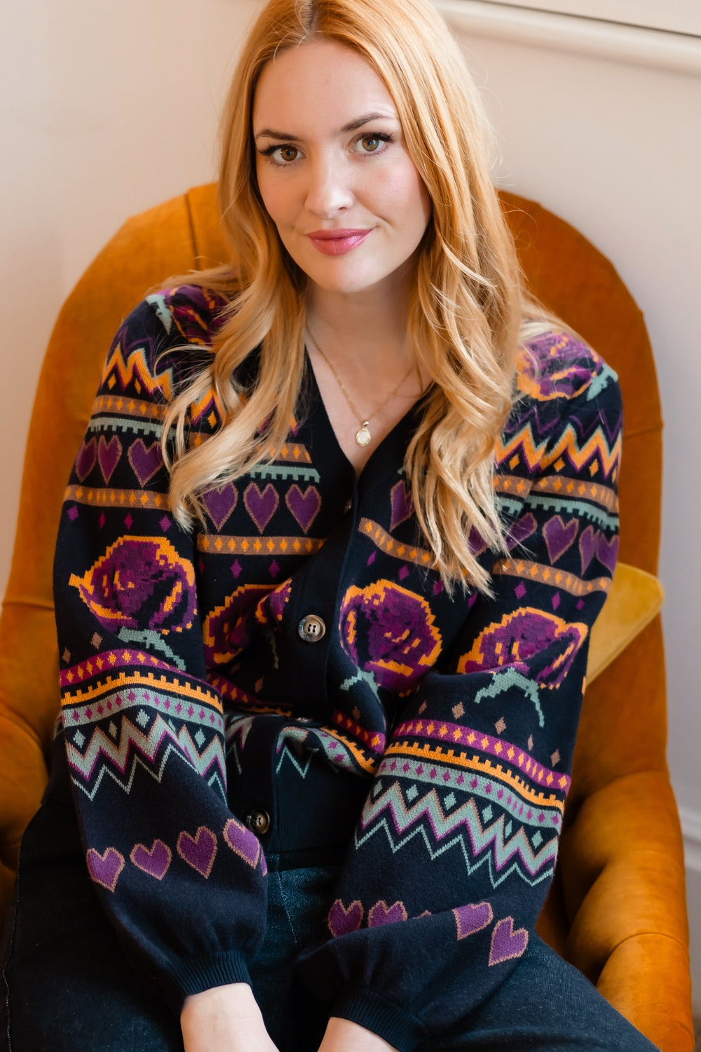 A woman with blonde hair sits in a mustard yellow armchair wearing a dark navy cardigan sweater with a colorful pattern of purple roses, hearts, and geometric stripes.