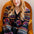 A woman with blonde hair sits in a mustard yellow armchair wearing a dark navy cardigan sweater with a colorful pattern of purple roses, hearts, and geometric stripes.