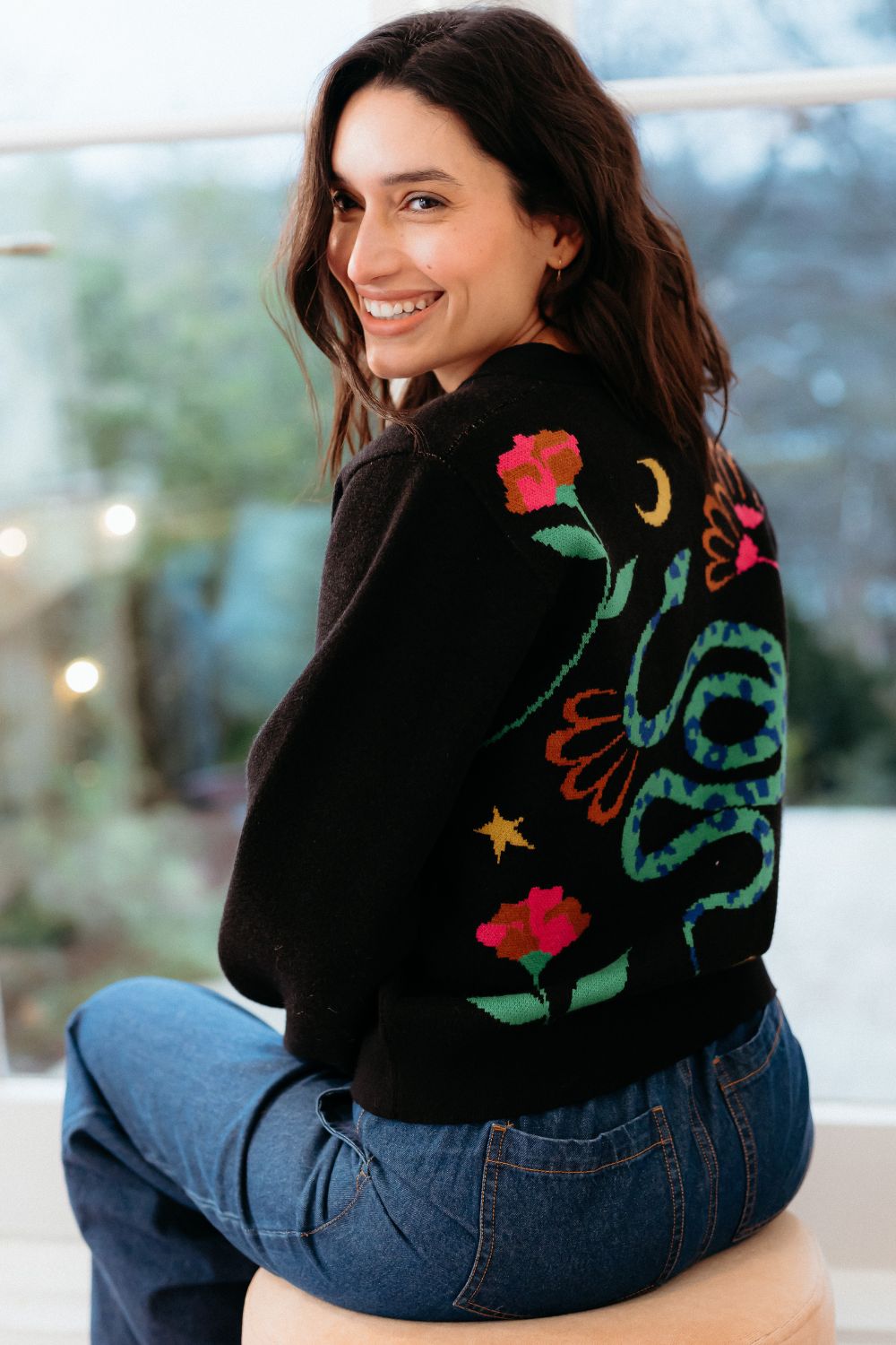Woman wearing a black cardigan with colourful floral embroidery sitting on a stool.