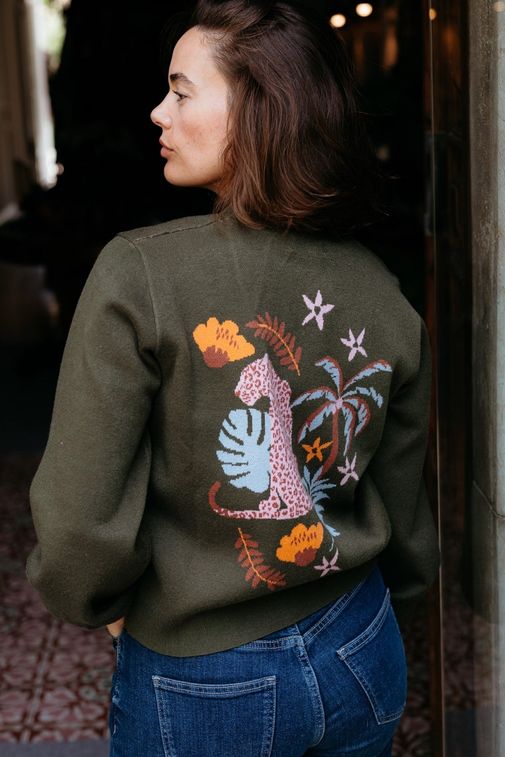 Person wearing a green sweatshirt with colorful designs on the back, standing indoors.