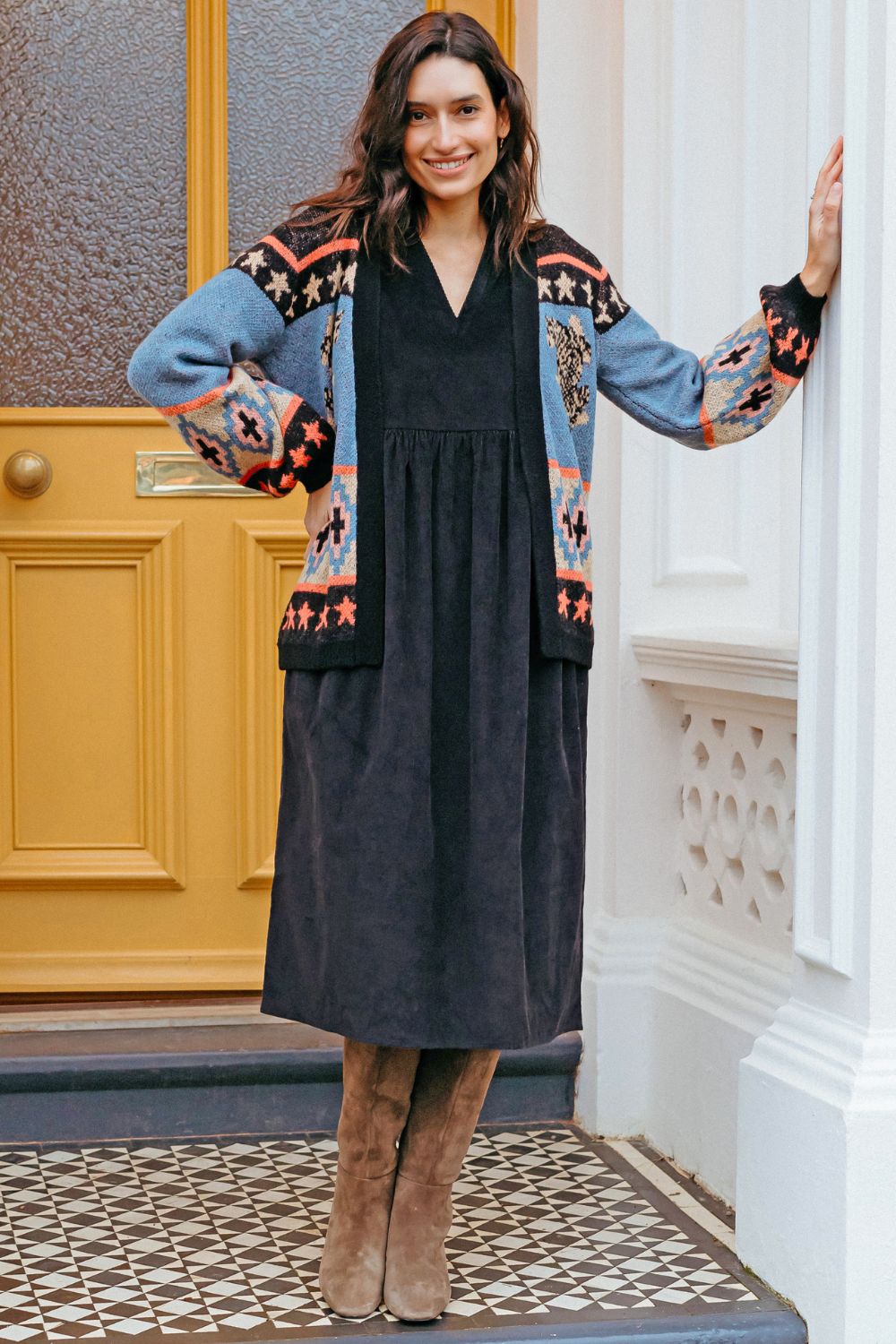 Woman wearing a black dress and patterned cardigan standing in front of a door.