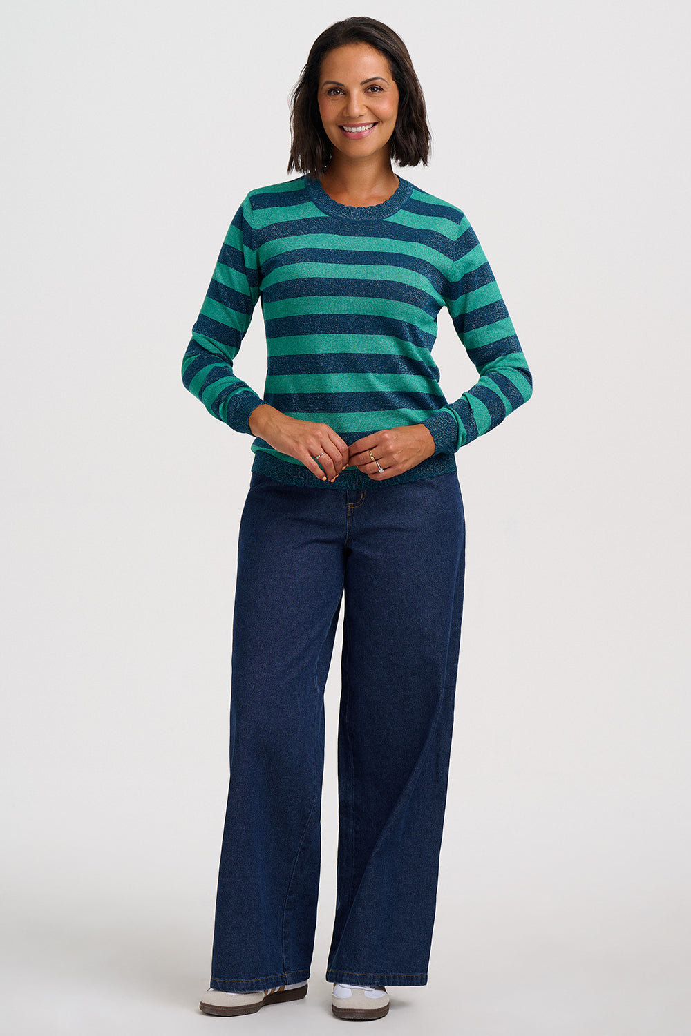 A woman modeling a green and navy striped long-sleeve knit top with dark blue wide-leg jeans, standing against a light gray studio background.