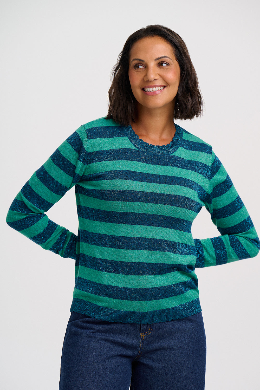 A woman wearing a long-sleeve crewneck sweater with horizontal stripes in teal green and dark navy blue, paired with dark blue jeans, against a light gray studio background.