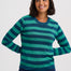 A woman wearing a long-sleeve crewneck sweater with horizontal stripes in teal green and dark navy blue, paired with dark blue jeans, against a light gray studio background.