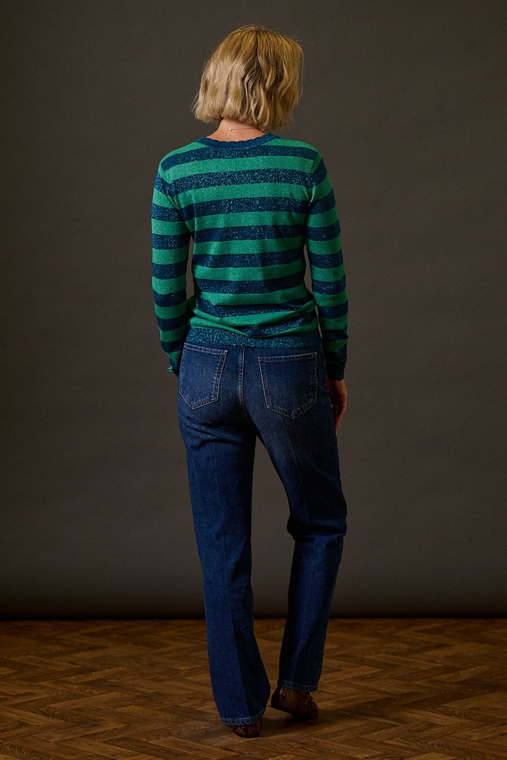 Person wearing a green and blue striped sweater and blue jeans against a brown background