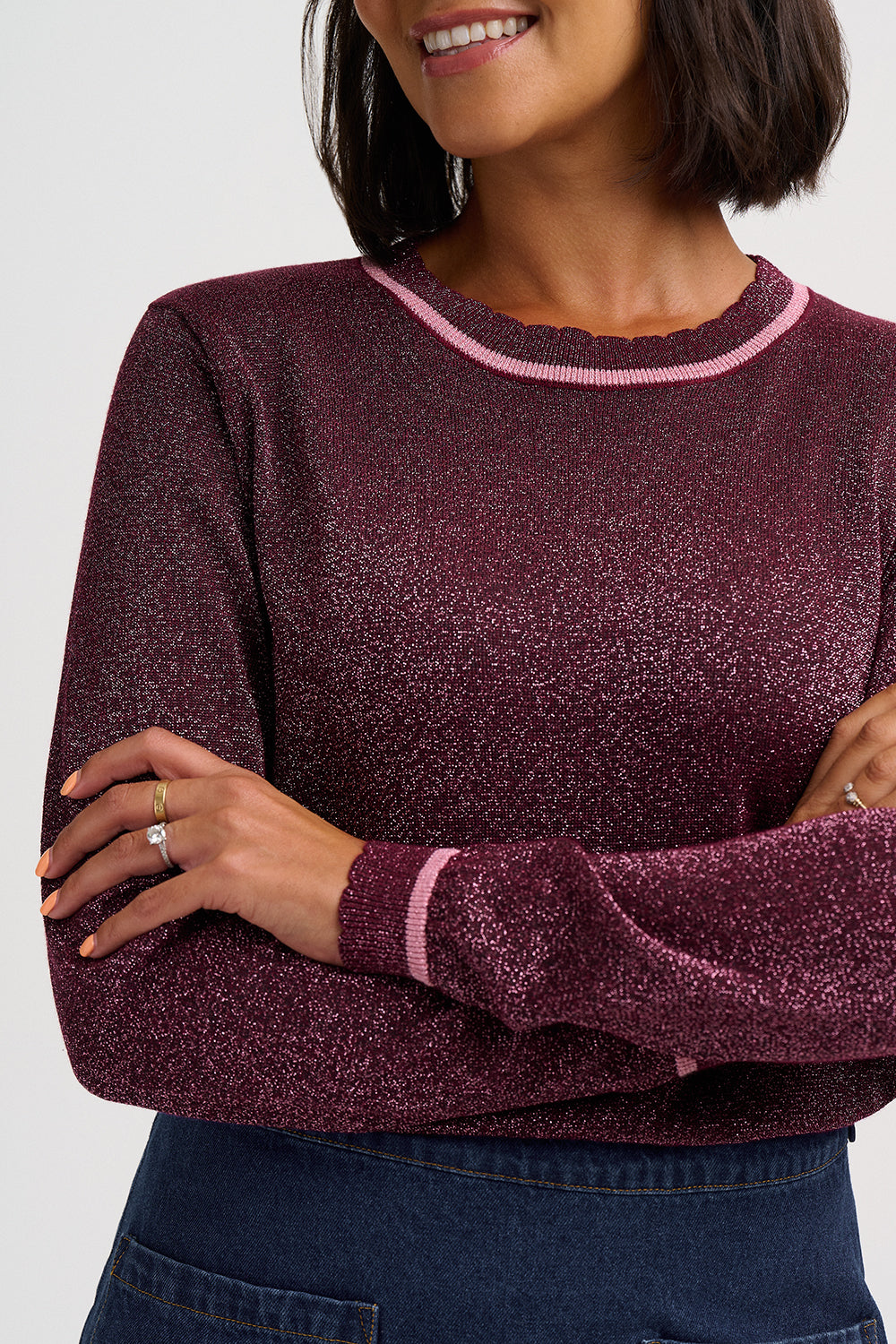 A close-up of a woman wearing a sparkling burgundy long-sleeve knit sweater with light pink trim on the crew neck and cuffs, paired with dark blue jeans.