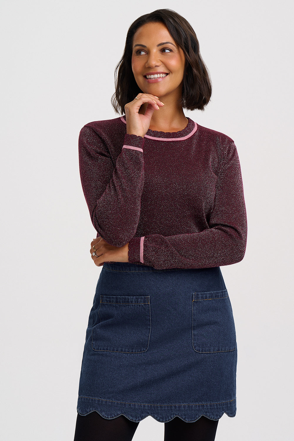 A woman modeling a maroon long-sleeve knit sweater with light pink trim on the crew neck and cuffs, paired with a dark blue denim skirt, against a light gray studio background.