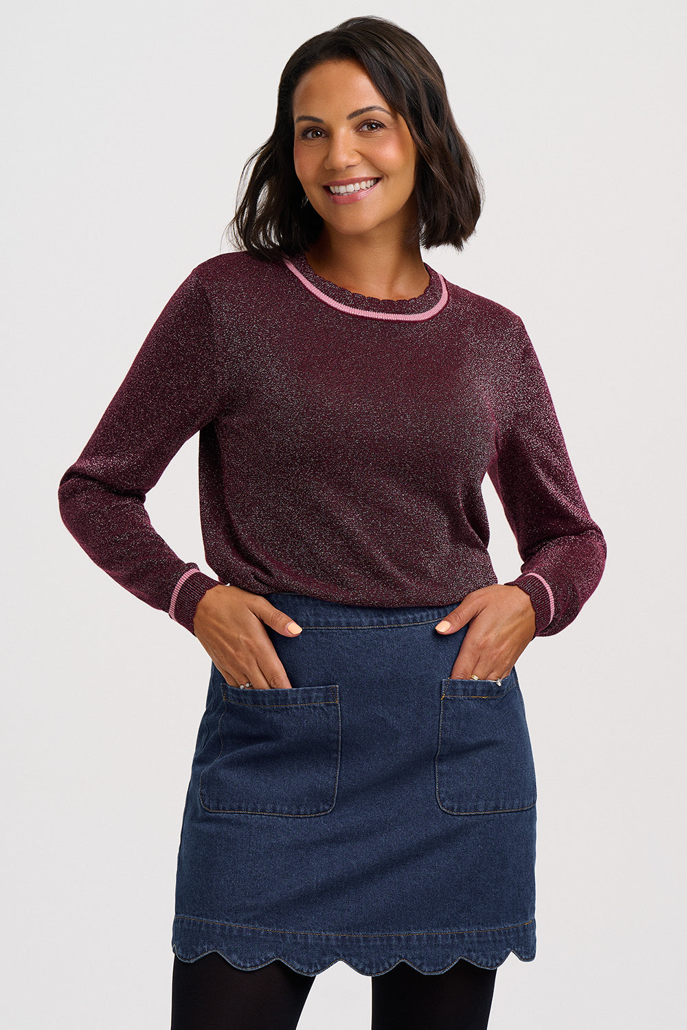 A woman modeling a long-sleeve knit sweater in a heathered burgundy color with light pink trim, paired with a dark blue denim skirt, against a light gray studio background.