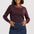 A woman modeling a long-sleeve knit sweater in a heathered burgundy color with light pink trim, paired with a dark blue denim skirt, against a light gray studio background.