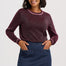 A woman modeling a long-sleeve knit sweater in a heathered burgundy color with light pink trim, paired with a dark blue denim skirt, against a light gray studio background.