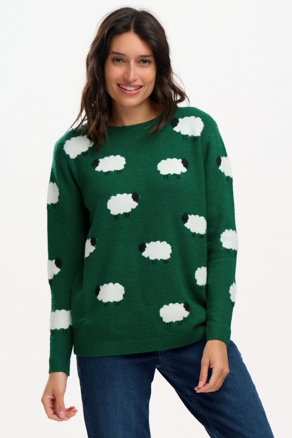 A woman wearing a dark green knit sweater with a white and black sheep pattern, paired with blue jeans, against a plain white background.