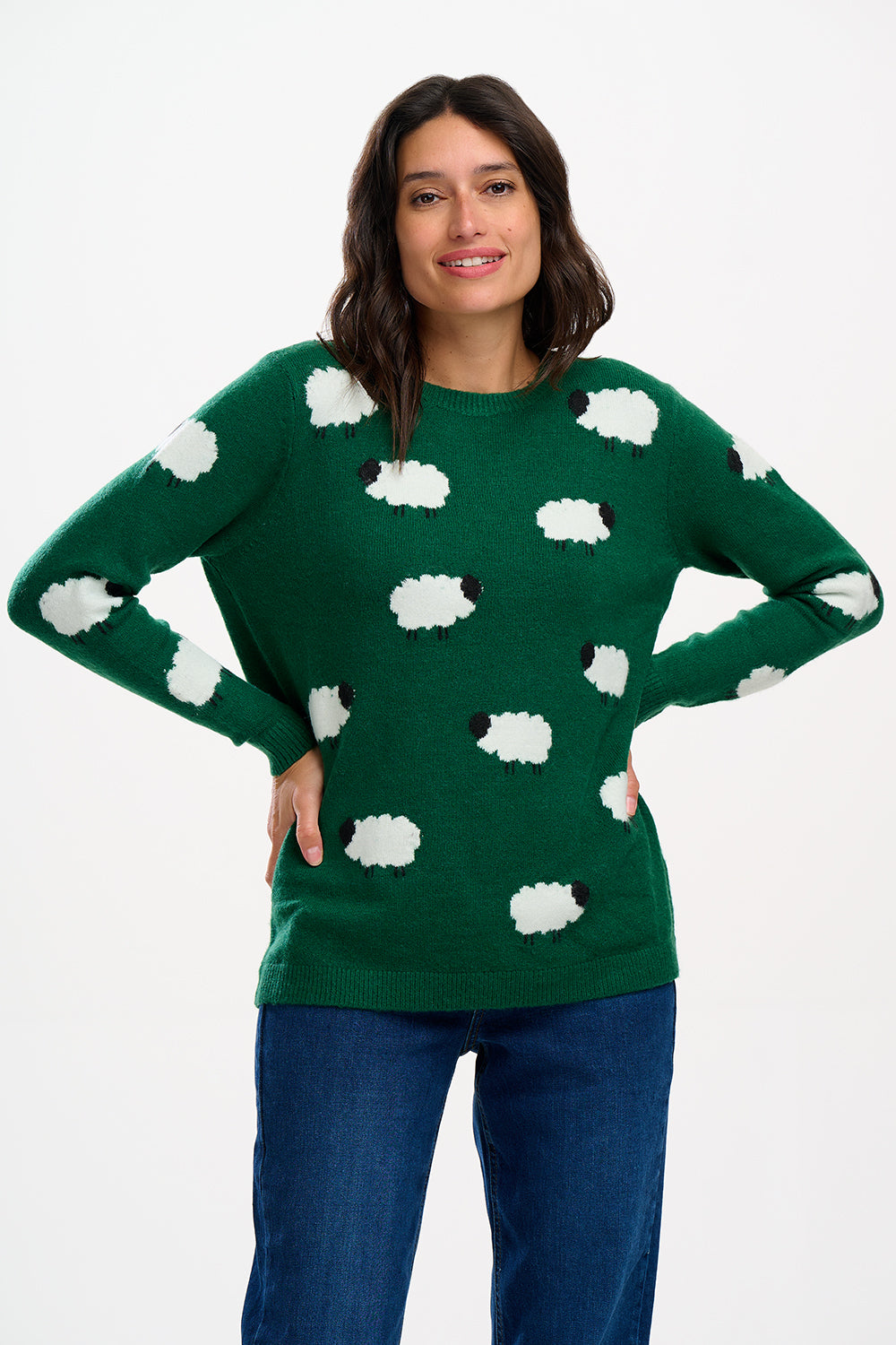 A woman modeling a green crewneck sweater with white sheep motifs, paired with blue jeans and white sneakers, against a plain white studio background.