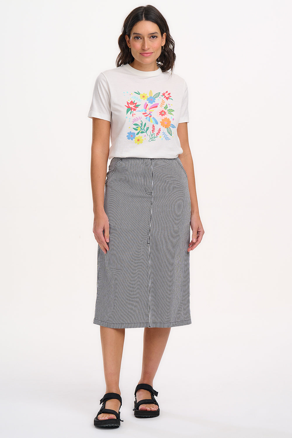 A woman wearing a fitted A-line midi skirt with monochrome stripes, paired with a white t-shirt featuring a colorful floral design, black sandals, and standing against a white background.