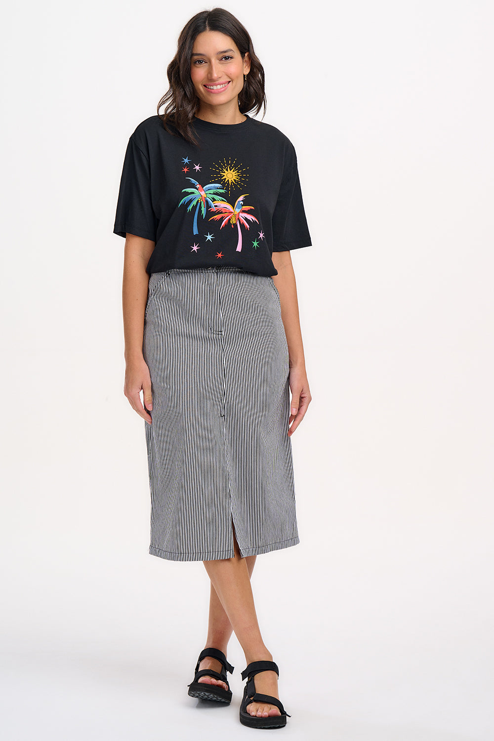 A woman wearing a fitted A-line midi skirt with monochrome stripes, paired with a white t-shirt featuring a colorful floral design, black sandals, and standing against a white background.