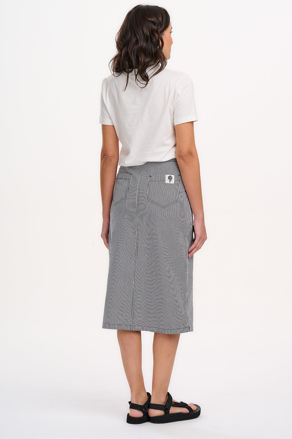 A woman wearing a fitted A-line midi skirt with monochrome stripes, paired with a white t-shirt featuring a colorful floral design, black sandals, and standing against a white background.