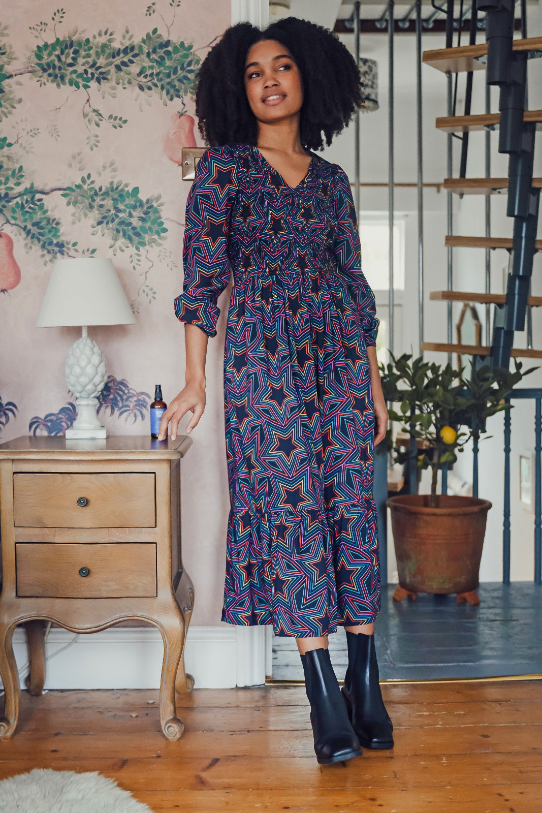 A woman standing in a room with vintage furniture, wearing a midi dress with a shirred elasticated bust and long sleeves, featuring a multicolored star print on a black base.