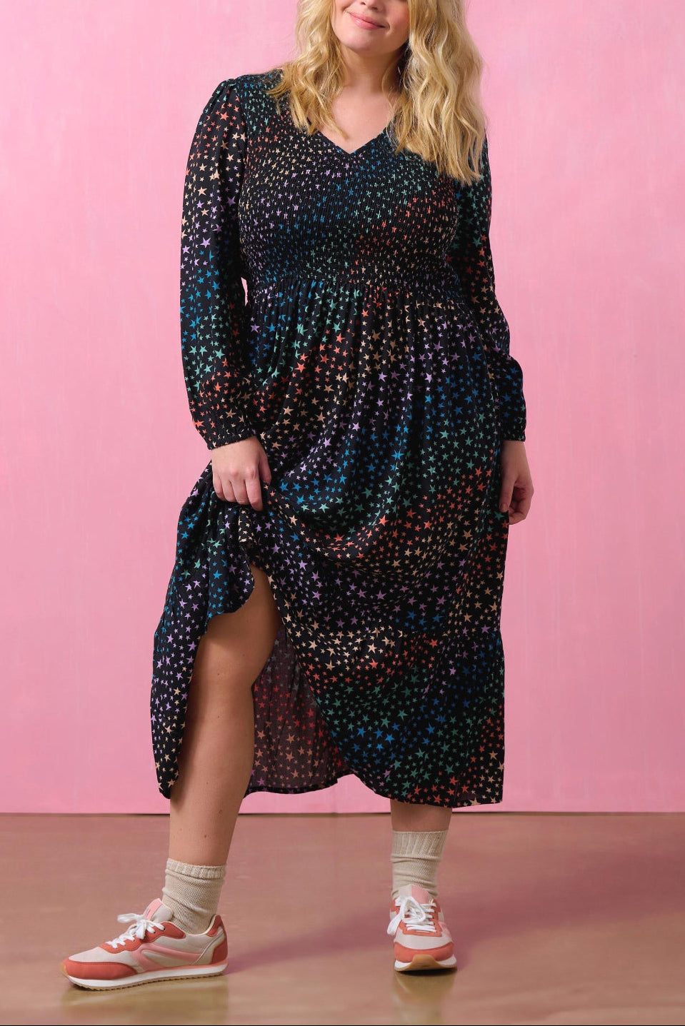 A woman standing in a studio setting, wearing an amberly shirred midi dress with a star waves print in black, with long blouson sleeves and an elasticated shirred bust.