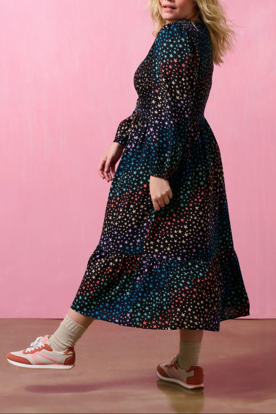 Woman wearing a long, dark dress with a colorful pattern against a pink background