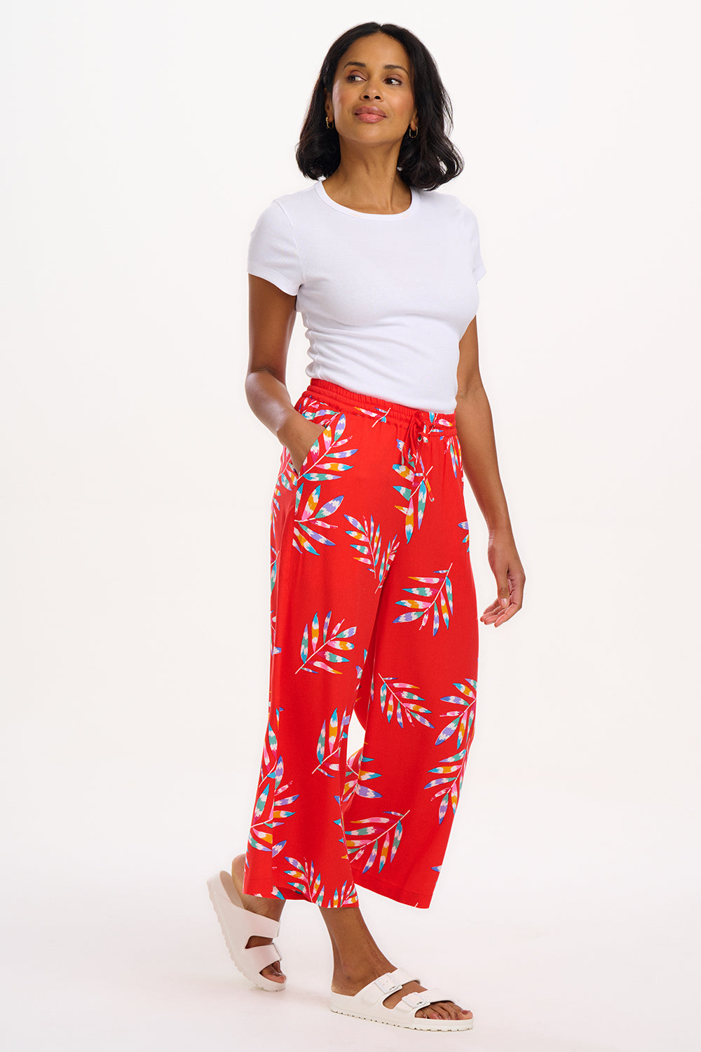 A woman modeling red cropped wide-leg trousers with a Rainbow Fronds print, paired with a white top and white sandals.