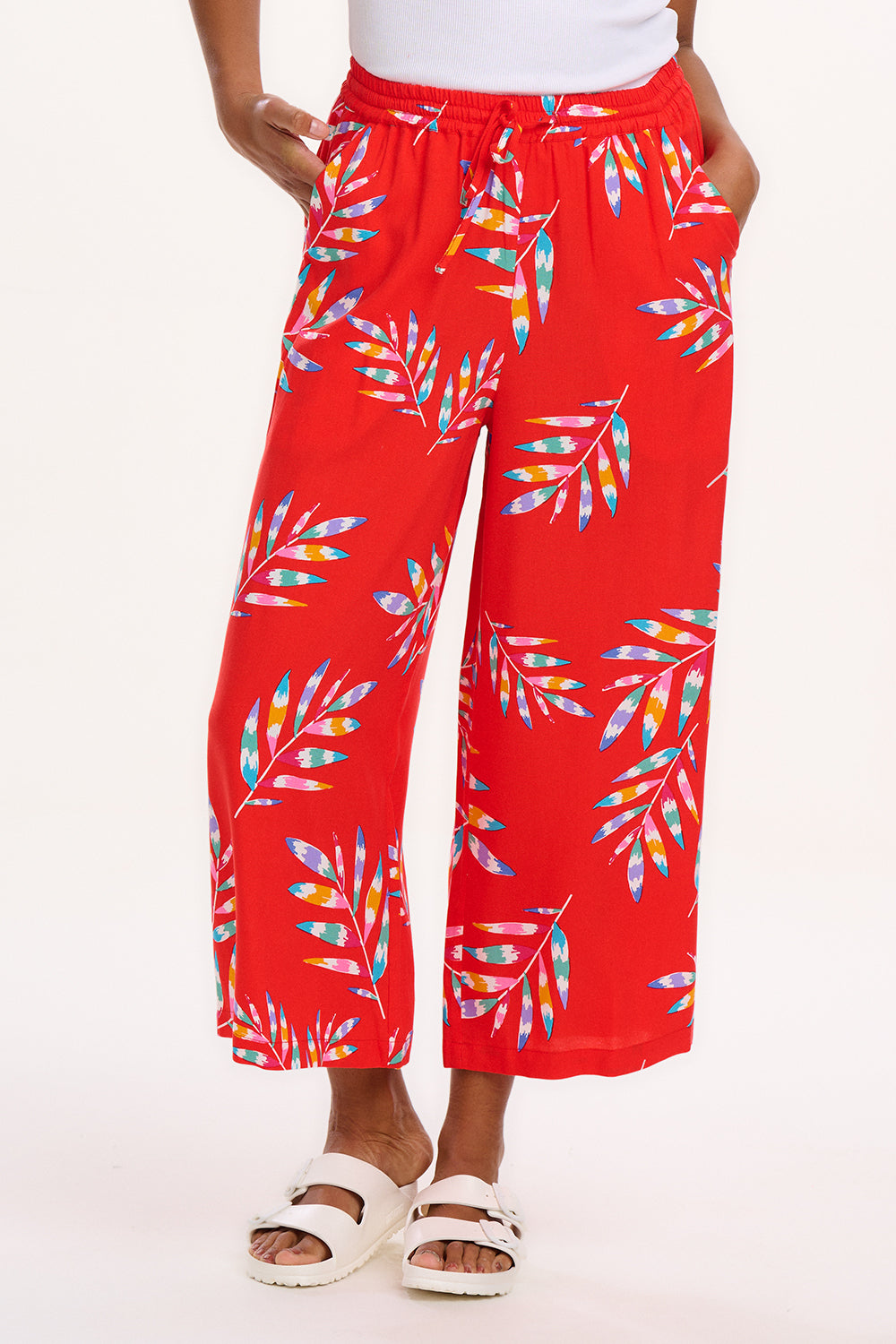 A model wearing red cropped wide-leg trousers with a colorful leaf pattern, standing against a white background.