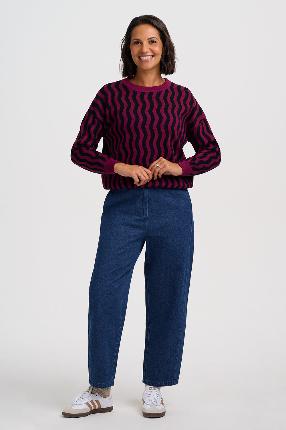 A woman modeling a dark purple and black wavy striped long-sleeve knit sweater, paired with dark blue jeans and white sneakers, against a light gray studio background.