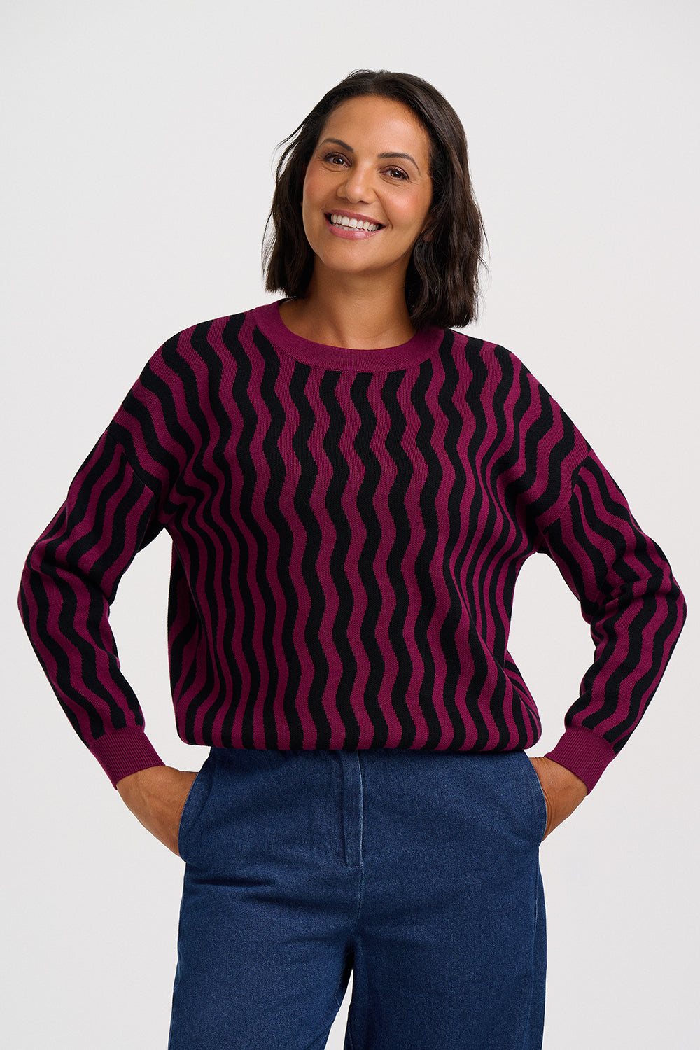 A woman wearing a long-sleeve crewneck sweater with a dark purple and black wavy stripe pattern, paired with blue jeans, against a light gray studio background.