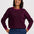 A woman wearing a long-sleeve crewneck sweater with a dark purple and black wavy stripe pattern, paired with blue jeans, against a light gray studio background.