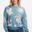 A woman wearing a blue and cream colored intarsia knit jumper with a tropical nights design, including white parrots, palm trees, stars, and suns on a pastel blue base.