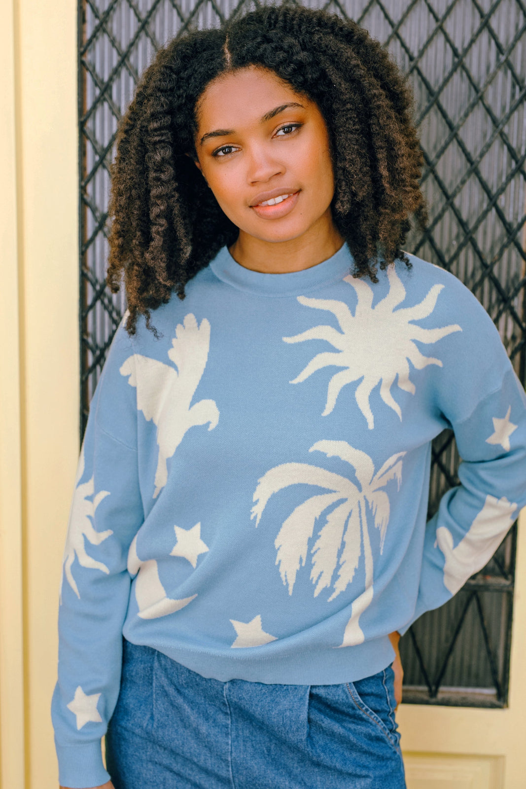 A woman wearing a blue and cream colored intarsia knit jumper with a tropical nights design, including white parrots, palm trees, stars, and suns on a pastel blue base.