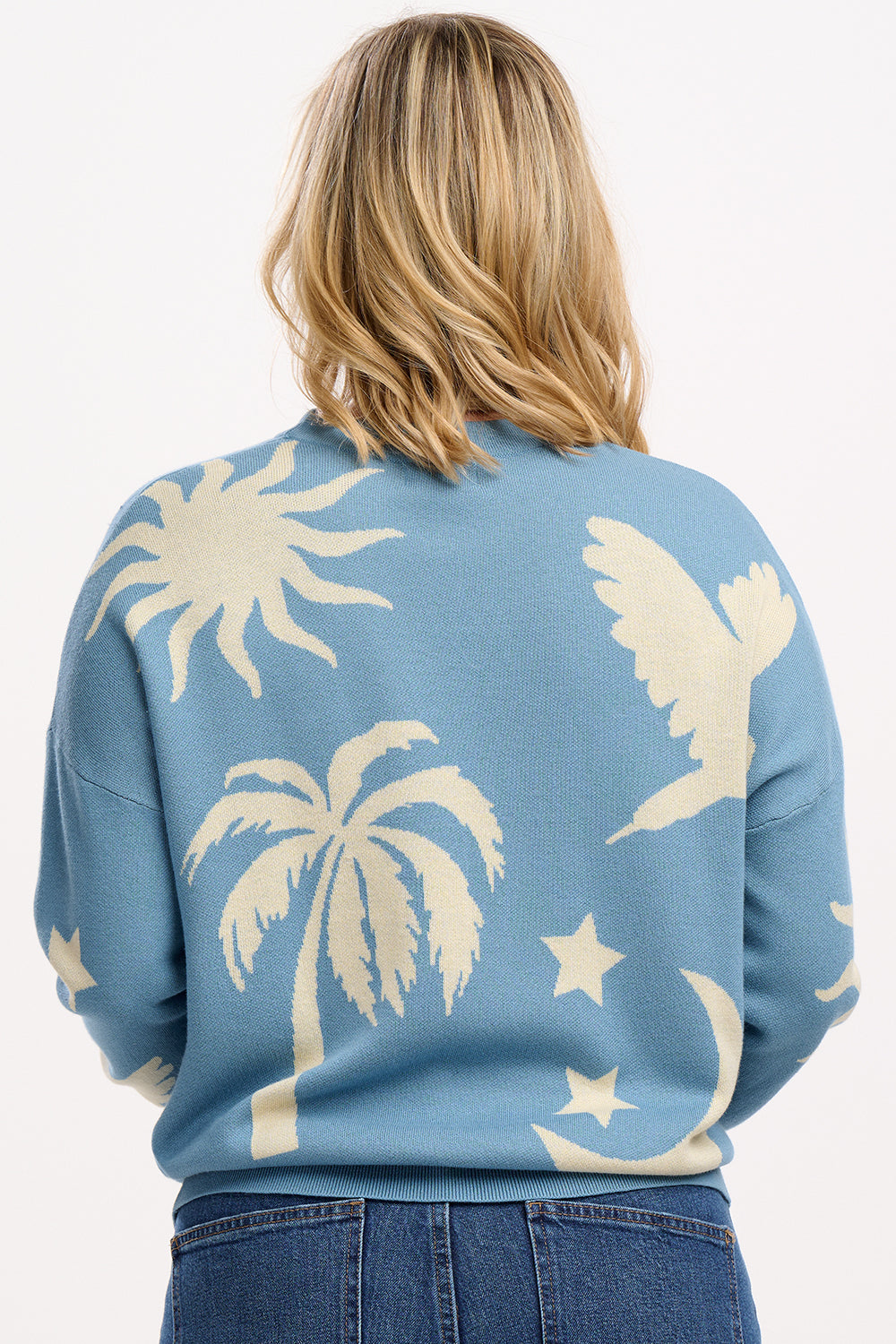 A woman wearing a blue and cream colored intarsia knit jumper with a tropical nights design, including white parrots, palm trees, stars, and suns on a pastel blue base.