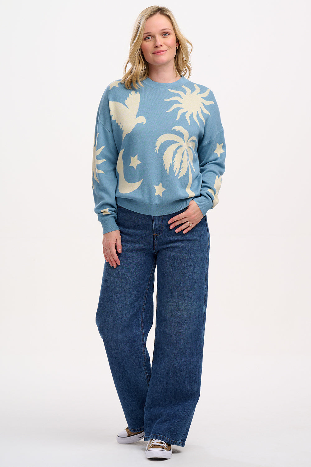 A woman wearing a blue and cream colored intarsia knit jumper with a tropical nights design, including white parrots, palm trees, stars, and suns on a pastel blue base.