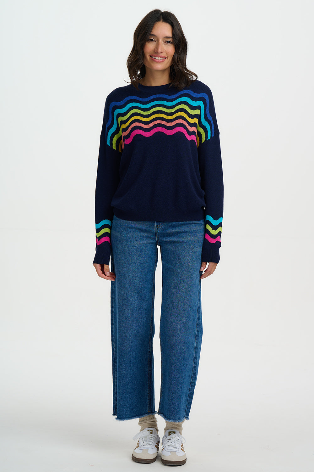 Woman wearing a navy sweater with colorful wave pattern and blue jeans on a white background
