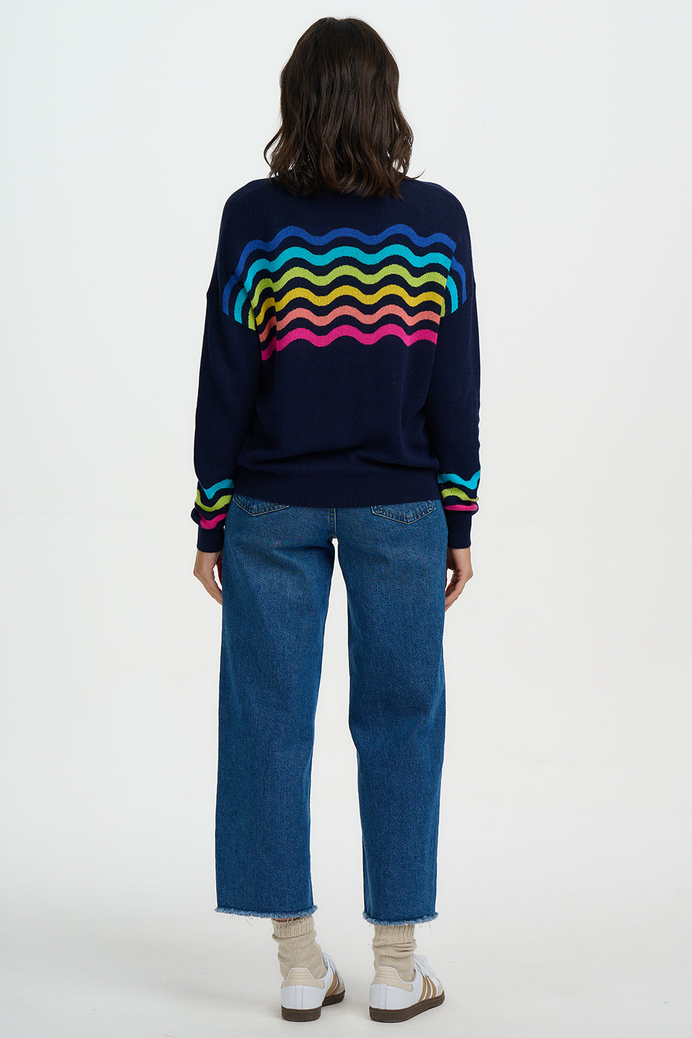 Person wearing a navy sweater with colorful wavy pattern and blue jeans on a white background