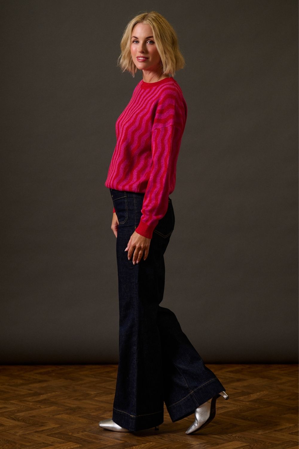 Woman wearing a pink sweater and dark blue flared pants against a plain background