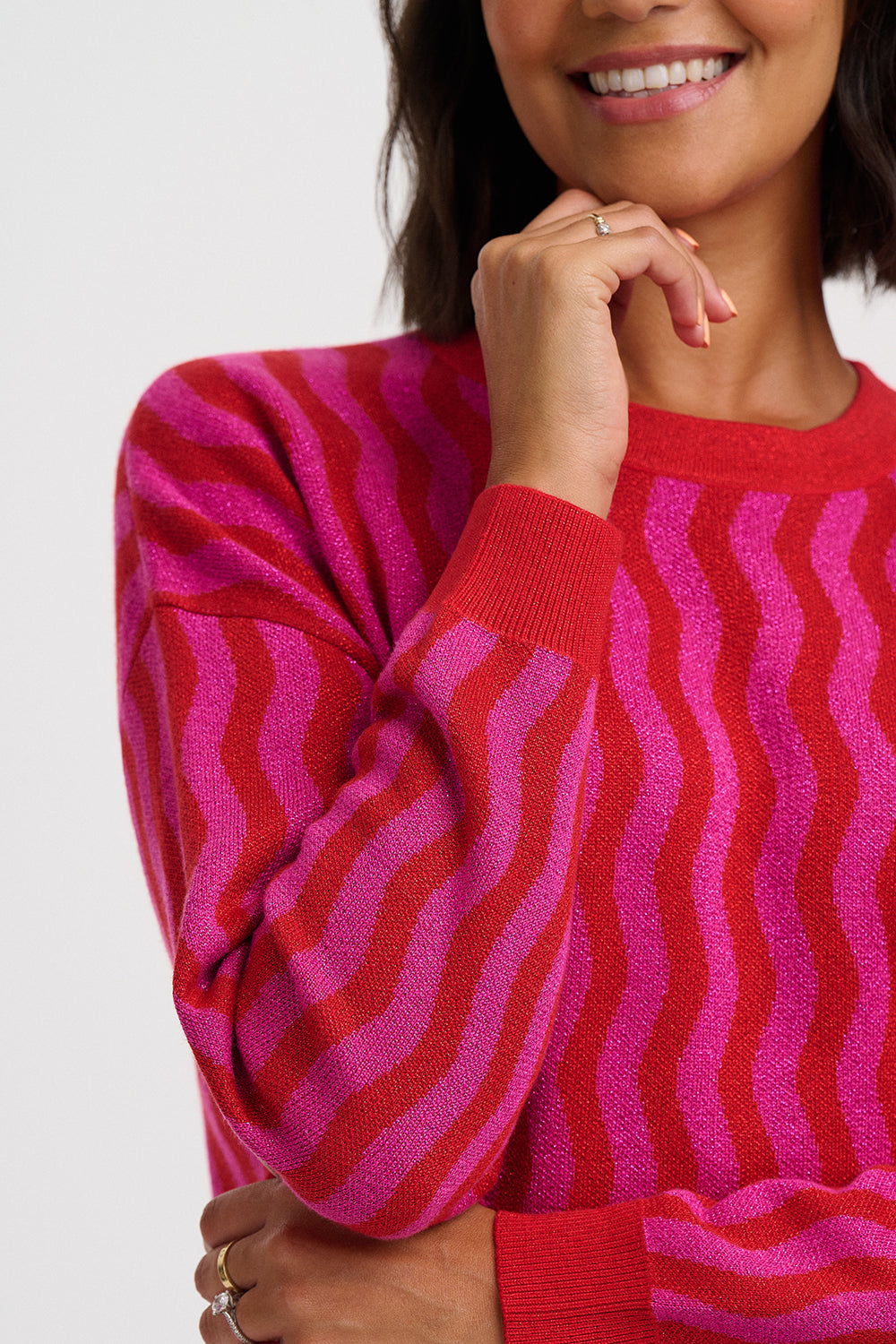 A close-up of a woman wearing a bright pink and red wavy striped knit sweater against a light gray studio background.