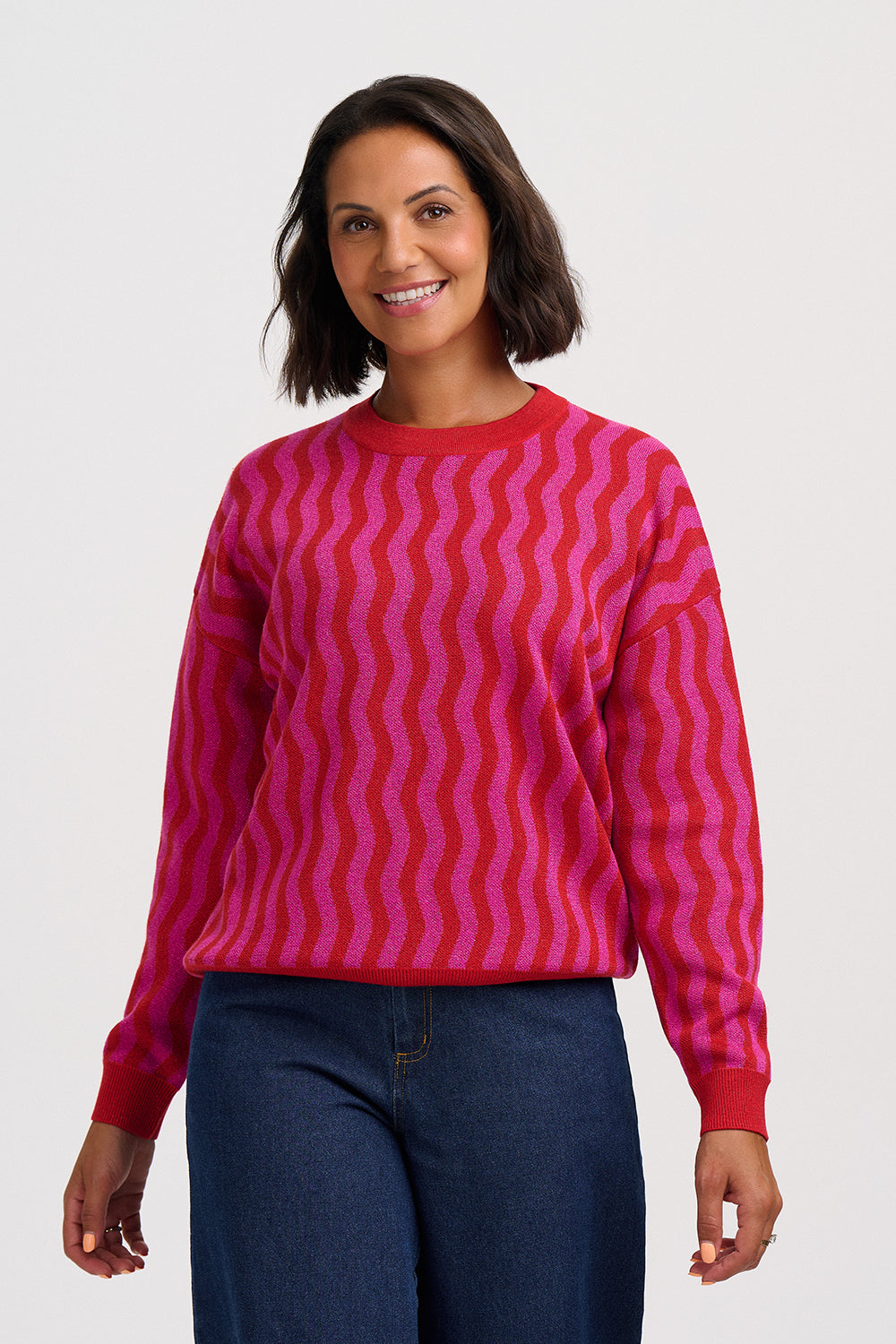 A woman wearing a bright pink and magenta wavy striped knit sweater with dark blue jeans, standing against a light gray studio background.