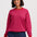 A woman wearing a bright pink and magenta wavy striped knit sweater with dark blue jeans, standing against a light gray studio background.
