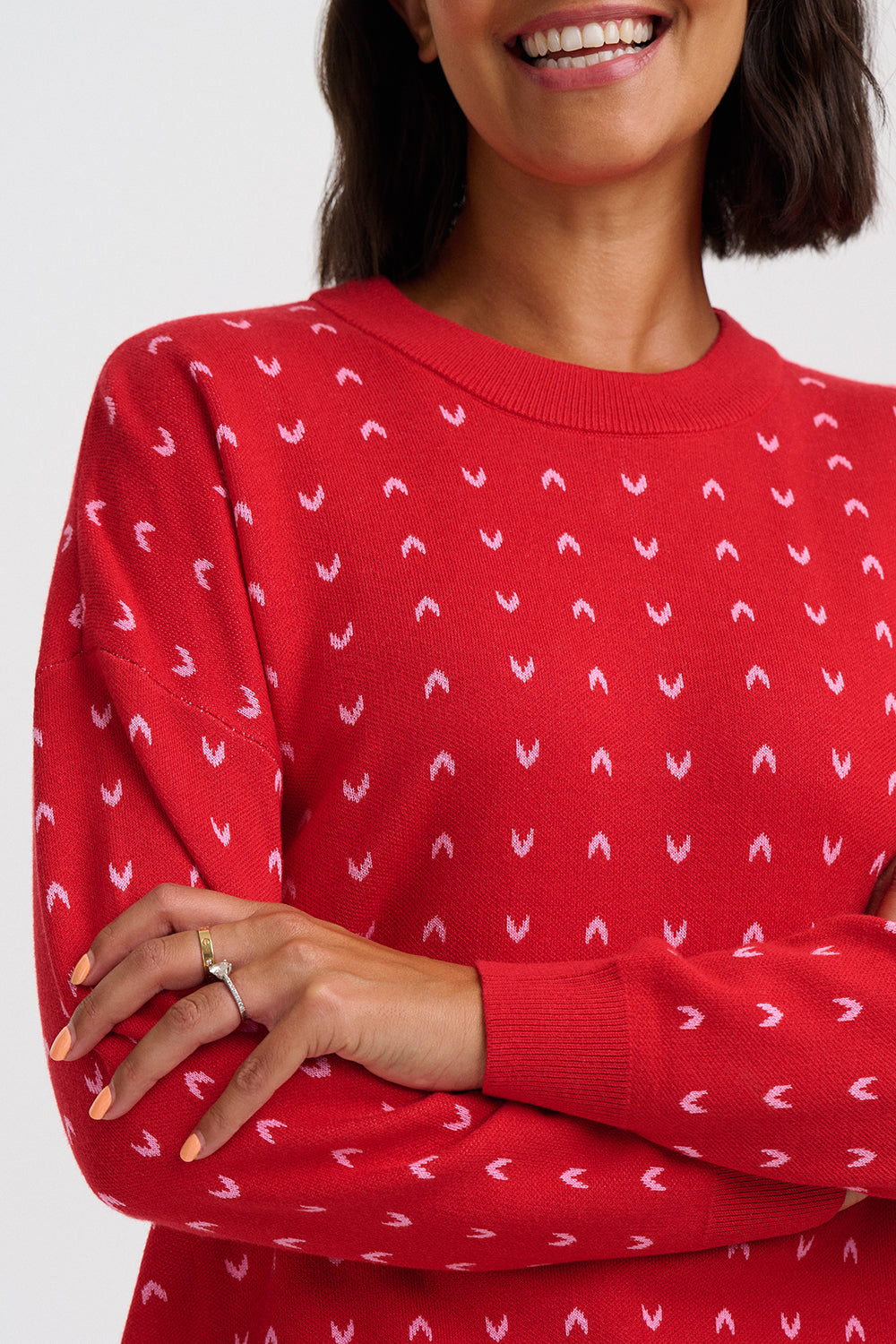 A close-up of a woman wearing a red crewneck sweater with a repeating white chevron pattern, shown from the chest up against a light gray background.