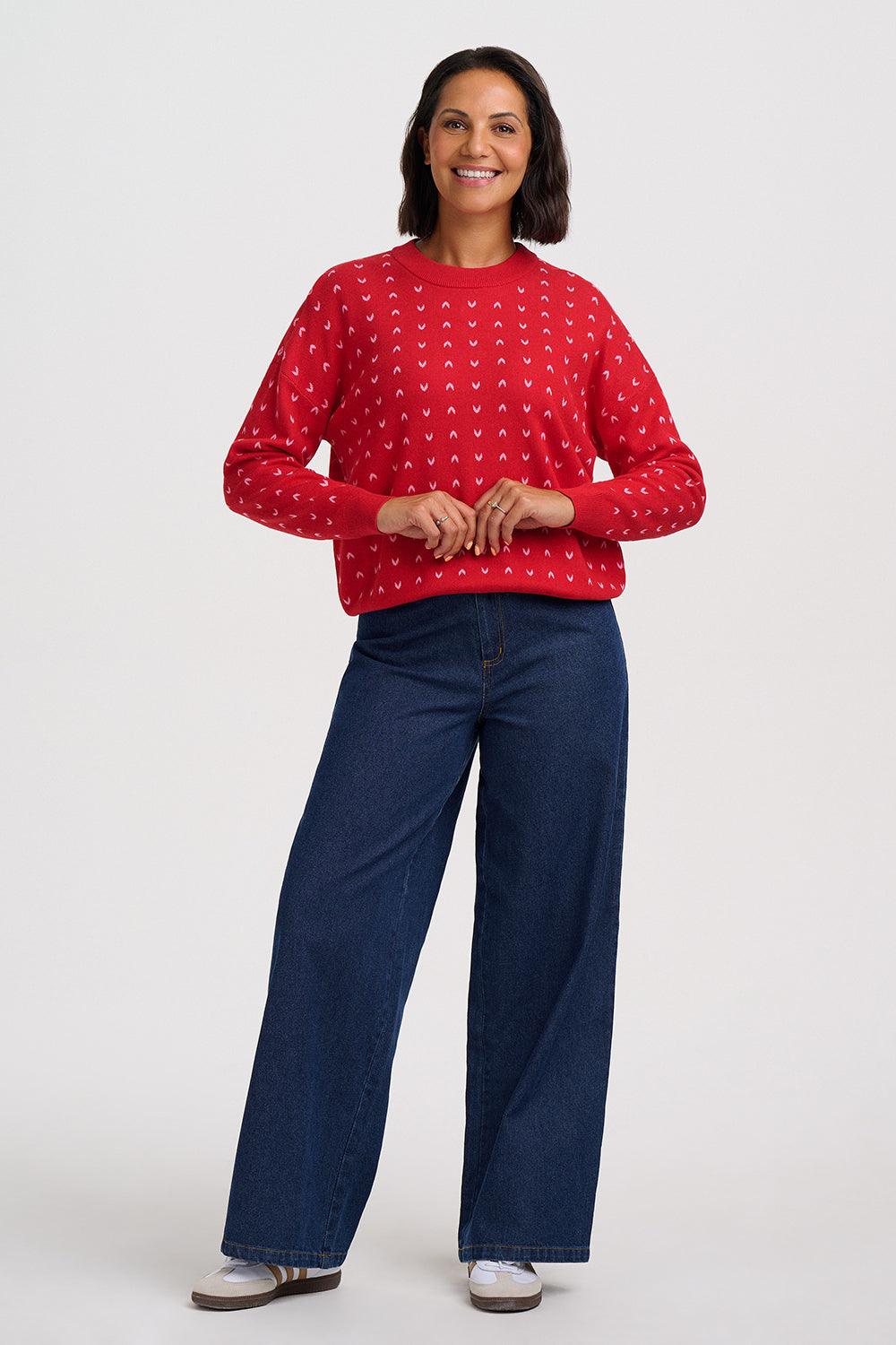 A woman modeling a red crewneck sweater with a white repeating pattern, paired with dark blue wide-leg jeans and white sneakers, against a light gray studio background.