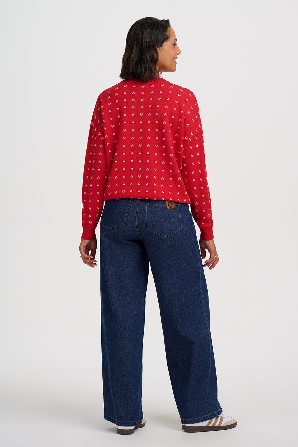 A woman shown from the back wearing a red long-sleeve knit top with a small repeating white pattern, paired with dark blue wide-leg jeans and white sneakers, against a plain light gray studio background.
