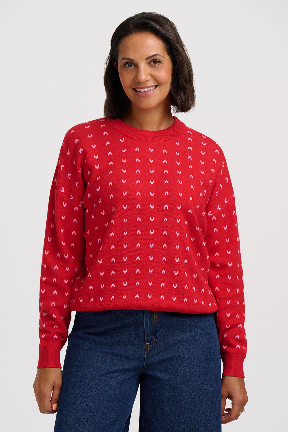 A woman wearing a red crewneck sweatshirt with a repeating white pattern, paired with dark blue jeans, against a light gray studio background.