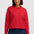 A woman wearing a red crewneck sweatshirt with a repeating white pattern, paired with dark blue jeans, against a light gray studio background.