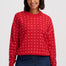 A woman wearing a red crewneck sweatshirt with a repeating white pattern, paired with dark blue jeans, against a light gray studio background.