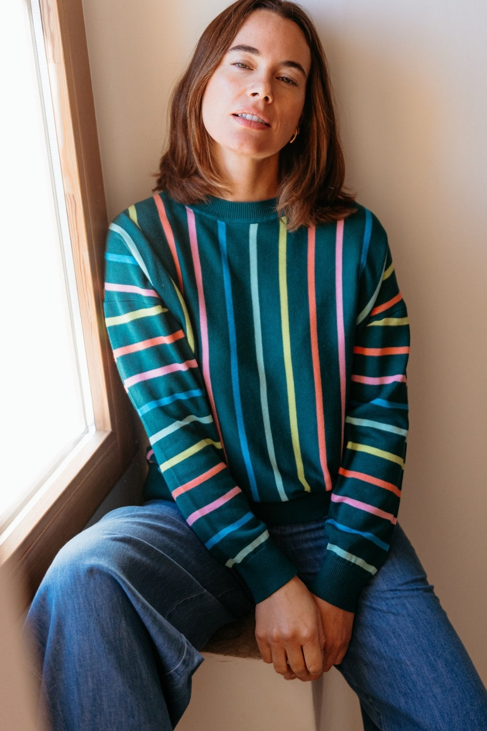 Woman wearing a colorful striped sweater sitting by a window.