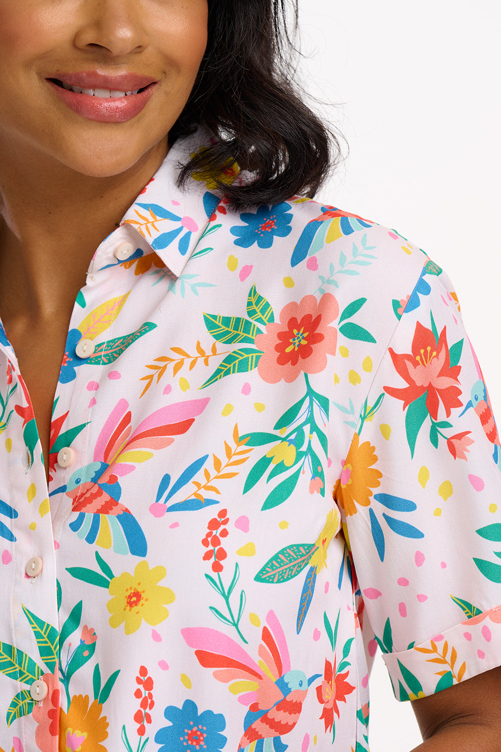 A woman wearing an off-white shirt with a tropical hummingbird print, short sleeves, and rolled-up cuffs.
